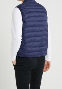 Námořnické modré lehké péřové vesty s vodorovným prošíváním, stojáčkovým límcem a kapsami na zip, nošené přes bílou košili s dlouhým rukávem.