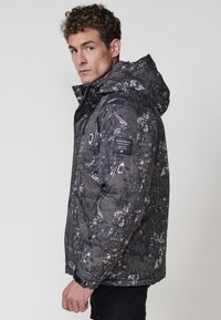 Chaqueta puffer de camuflaje gris con acabado texturizado y capucha. Presenta un parche con el logo en el brazo izquierdo y puños ajustables.