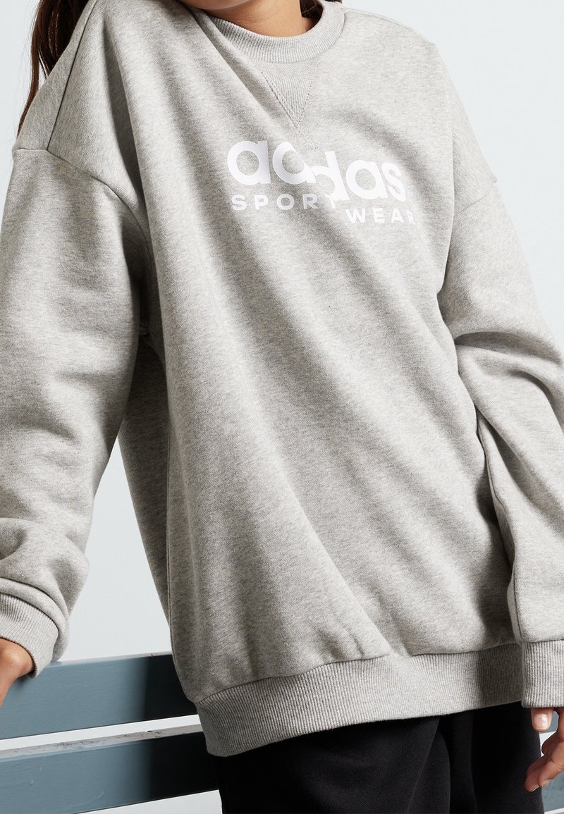 Šedá mikina vyrobená z měkkého materiálu s kulatým výstřihem, spadlými rameny, žebrovanými manžetami a výrazným bílým logem "adidas SPORT WEAR".