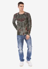 Langarm, dunkelgrünes Grafik-T-Shirt mit roten und schwarzen Prints; kombiniert mit hellblauen, verwaschenen Jeans und weißen Sneakern.