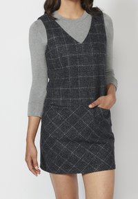 Navy plaid dress with a deep V-neckline and a fitted silhouette, layered over a light gray long-sleeve shirt. Textured fabric, two pockets.