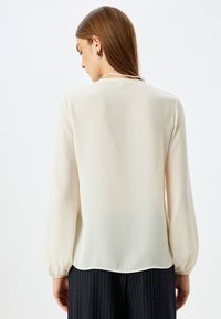 Lys beige, langærmet bluse i et glat, gennemsigtigt stof med rund halsudskæring og samlede manchetter, parret med mørke stribede bukser.