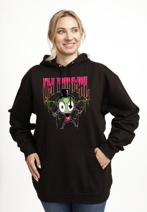 Uśmiechnięta kobieta w czarnej bluzie z kapturem z kolorową postacią z kreskówki i napisem „I'M NORMAL”, stojąca z rękami w przednich kieszeniach.