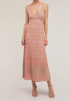 Vrouw draagt een mouwloze midi-jurk met V-hals in roze met textuurpatronen, gecombineerd met open-teen enkellaarsjes met enkelbandje.
