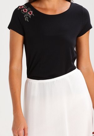 Haut noir à manches courtes avec broderie florale près de l'épaule, associé à une jupe plissée blanche. Design simple et ajusté.