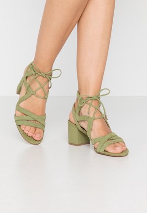 Las sandalias de tacón alto de ante verde cuentan con múltiples correas cruzadas, ataduras al tobillo y un tacón cuadrado, combinando textura y estabilidad.