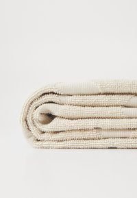 Asciugamano beige con texture, arrotolato ordinatamente, realizzato in un morbido tessuto intrecciato con un motivo sottile e un bordo leggermente più spesso, posizionato su uno sfondo bianco.
