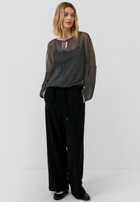 Vrouw in een doorzichtige sprankelende zwarte blouse met striknek over een zwarte camisole, met losse zwarte broeken en zwarte flats, staand tegen een witte achtergrond.