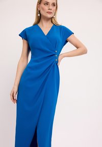 Niebieska sukienka wrap z krótkimi rękawkami, z drapowaną strukturą i dekoracyjnymi złotymi guzikami po boku. Posiada rozcięcie z boku dla dodatkowego akcentu.