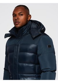 Hombre vestido con una chaqueta de invierno acolchada en azul marino, con un alto cuello y capucha, mirando ligeramente de lado frente a un fondo liso.