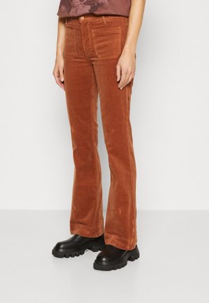 Osoba stojící před jednobarevným pozadím, oblečená v rezavě hnědých manšestrových kalhotách s předními našitými kapsami a černých botách s výraznou podrážkou.