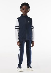 Granatowy dres z białymi akcentami. Kurtka zapinana na zamek z paskami na rękawach; spodnie mają boczne paski. Wykonany z gładkiego materiału; casualowy fason.