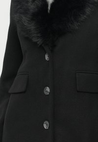 Cappotto in lana nera con colletto in pelliccia sintetica, chiusura a bottoni e due tasche frontali, leggermente testurizzato e dal design sartoriale.
