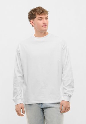 Młody mężczyzna o jasnobrązowych włosach, ubrany w prostą białą koszulę z długim rękawem i jasnoniebieskie dżinsy, patrzy w lewo.