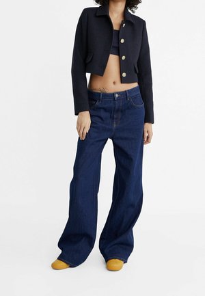 Woman wearing a cropped black buttoned jacket, wide-leg dark blue jeans, and mustard yellow shoes, standing against a plain white background.