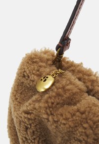 Sac en tissu marron duveteux avec une sangle en cuir marron et un charm en métal doré attaché près du fermoir de la poignée.