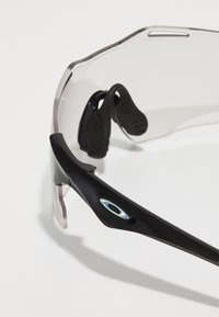 Czarne sportowe okulary przeciwsłoneczne z przezroczystymi soczewkami, zakrzywioną konstrukcją oprawki, gumowymi noskami oraz charakterystycznym logo na zausznicy.