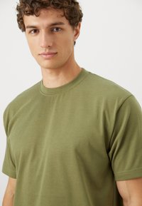 Olivgrünes Baumwoll-T-Shirt mit Rundhalsausschnitt. Kurze Ärmel und glatte Textur, passt eng am Körper an, ohne Verzierungen.