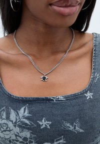 Colier din argint cu pandantiv în formă de inimă de culoare neagră, cu oase încrucișate, purtat peste un top fără mâneci cu model floral gri.