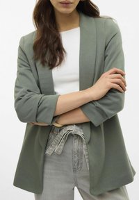 Grijs-groene blazer met een ontspannen fit, open voorkant en opgerolde mouwen, gecombineerd met een witte top en grijze hoogsluitende broek.