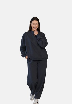 Kobieta z długimi ciemnymi włosami, uśmiechająca się, ubrana w ciemną, oversize'ową bluzę zapinaną na zamek oraz pasujące spodnie, z jedną ręką przy brodzie.