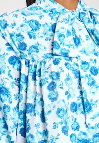 Blusa floral em branco e azul-claro. Apresenta um decote com laço, fitas na cintura e um padrão de flores de rosa em diferentes tons de azul.