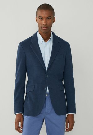 Marineblaues Blazer aus strukturiertem Stoff, zwei Vorderentaschen, Schößchen mit Revers und zwei Knopfverschluss, kombiniert mit einem hellblauen Hemd mit Knopfleiste.
