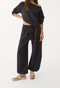 Zwarte off-the-shoulder sweatshirt met lange mouwen, gecombineerd met zwarte joggers met elastische taille. Bruine en beige sneakers maken de outfit compleet.