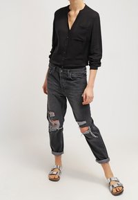 Person wearing black buttoned long-sleeve shirt, cuffed ripped dark gray jeans, and silver open-toe sandals standing on plain background.