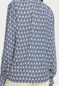 Blusa a maniche lunghe di colore blu navy con un motivo a foglie bianche. Presenta polsini arricciati e un tessuto morbido e strutturato. Il design posteriore è semplice e senza cuciture.