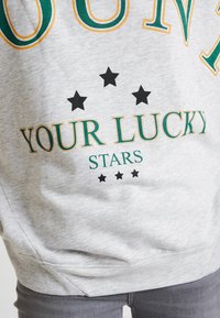 Grå sweatshirt med grön och orange text där det står "YOUR LUCKY STARS" och fem svarta stjärnor ovanför texten. Mjuk textur.