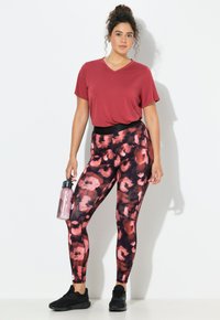 Rode v-hals t-shirt gecombineerd met zwart-roze geblokte legging. Model houdt een doorzichtige waterfles vast en draagt zwarte sportschoenen.