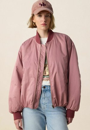 Jeune femme aux cheveux blonds courts portant une veste bomber rose poudré, un jean bleu clair, un t-shirt graphique, des boucles d'oreilles créoles dorées et une casquette rose assortie.