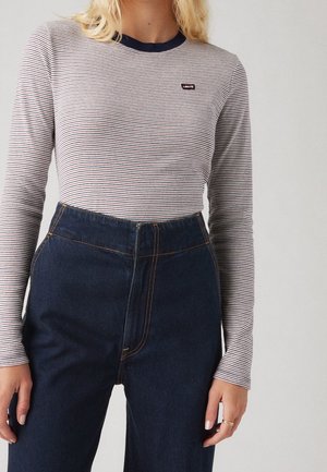 Woman wearing a fitted long-sleeve white and navy striped shirt with a small chest logo, tucked into high-waisted dark blue jeans.