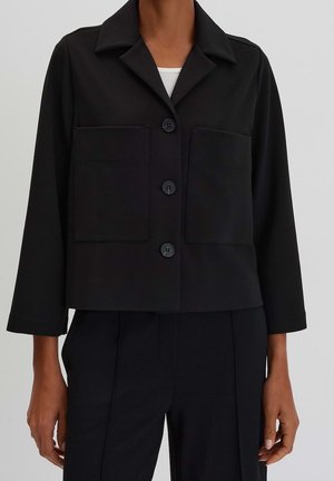 Nainen pukeutuneena mustaan lyhytmalliseen takkiin, jossa on kolme nappia, suuret etutaskut ja leveät hihat, ja mustat korkeavyötäröiset housut.