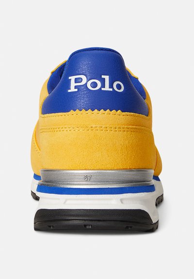 Żółty zamszowy sneaker z niebieskimi skórzanymi akcentami, białym logo i srebrnymi detalami na podeszwie. Posiada okrągły obcas i teksturowaną powierzchnię.