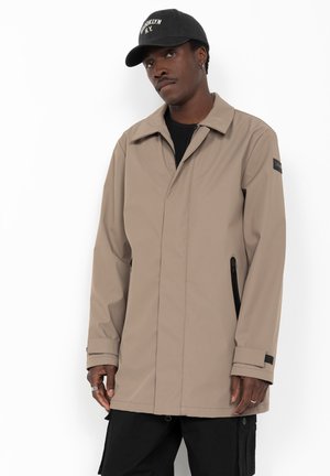 Homme portant une veste beige zippée et une casquette noire avec un texte blanc, debout devant un fond blanc