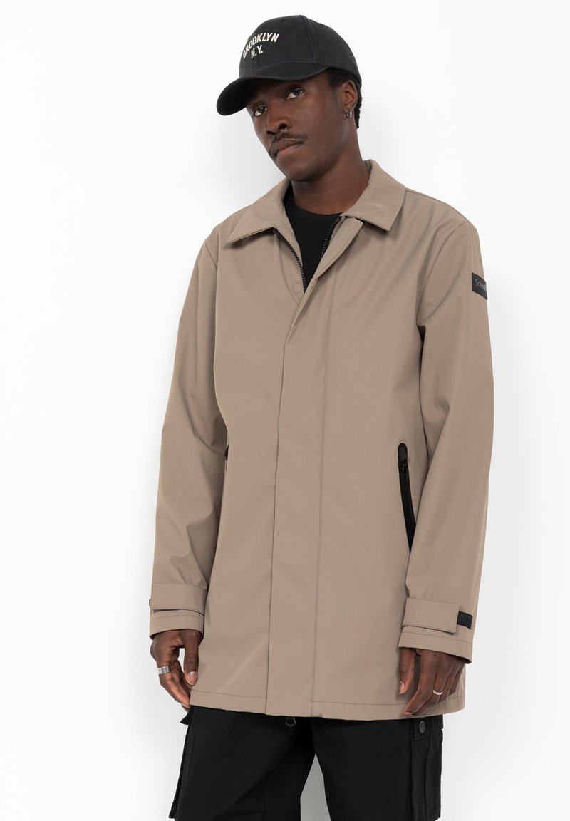 Homme portant une veste beige zippée et une casquette noire avec un texte blanc, debout devant un fond blanc