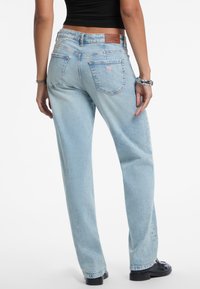 Mujer vestida con jeans rectos de Guess color azul claro y zapatos negros, vista desde la parte de atrás con las manos relajadas a los lados.