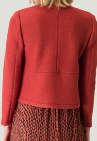 Červená texturovaná bunda s dlouhými rukávy, s přiléhavým střihem a viditelnými švy, doplněná vzorovanou sukní.