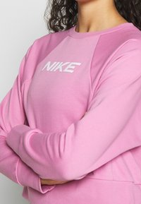 Personne aux cheveux bouclés portant un sweat-shirt rose Nike, bras croisés, visible du cou à la taille sur un fond uni.