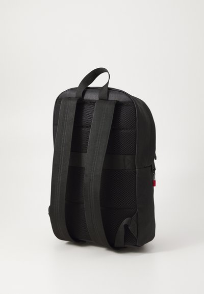 Sac à dos en tissu noir avec dos en maille rembourrée, bretelles réglables, poignée de transport et tirette de fermeture éclair rouge. Présente un design épuré et minimaliste.