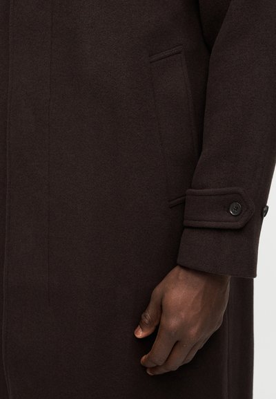 Μανίκι παλτού από σκουρόχρωμο καφέ μάλλινο ύφασμα με λεπτομέρεια κουμπωτού μανσετώ, πλαϊνή τσέπη και ένα σκουρόχρωμο χέρι που ακουμπά δίπλα στο παλτό.