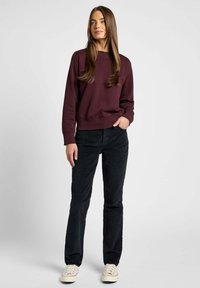 Bordowy sweatshirt z okrągłym dekoltem i prążkowanymi mankietami, w zestawieniu z czarnymi spodniami sztruksowymi. Model nosi kremowe sneakersy, stojąc na neutralnym tle.