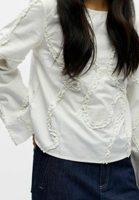 Hvit bomullsbluse med teksturerte volangkanter i et virvlende mønster, med lange ermer og en avslappet passform, kombinert med mørk denim.