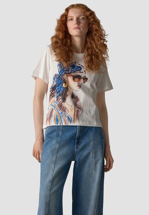 Woman with curly red hair wears a white t-shirt featuring a colorful illustrated woman with sunglasses and a blue headscarf, paired with loose denim jeans.