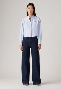 Femeie cu păr lung și închis la culoare, îmbrăcată cu o cămașă cu butoni de culoare albastru deschis și blugi largi de culoare albastru închis, stând pe un fundal simplu.