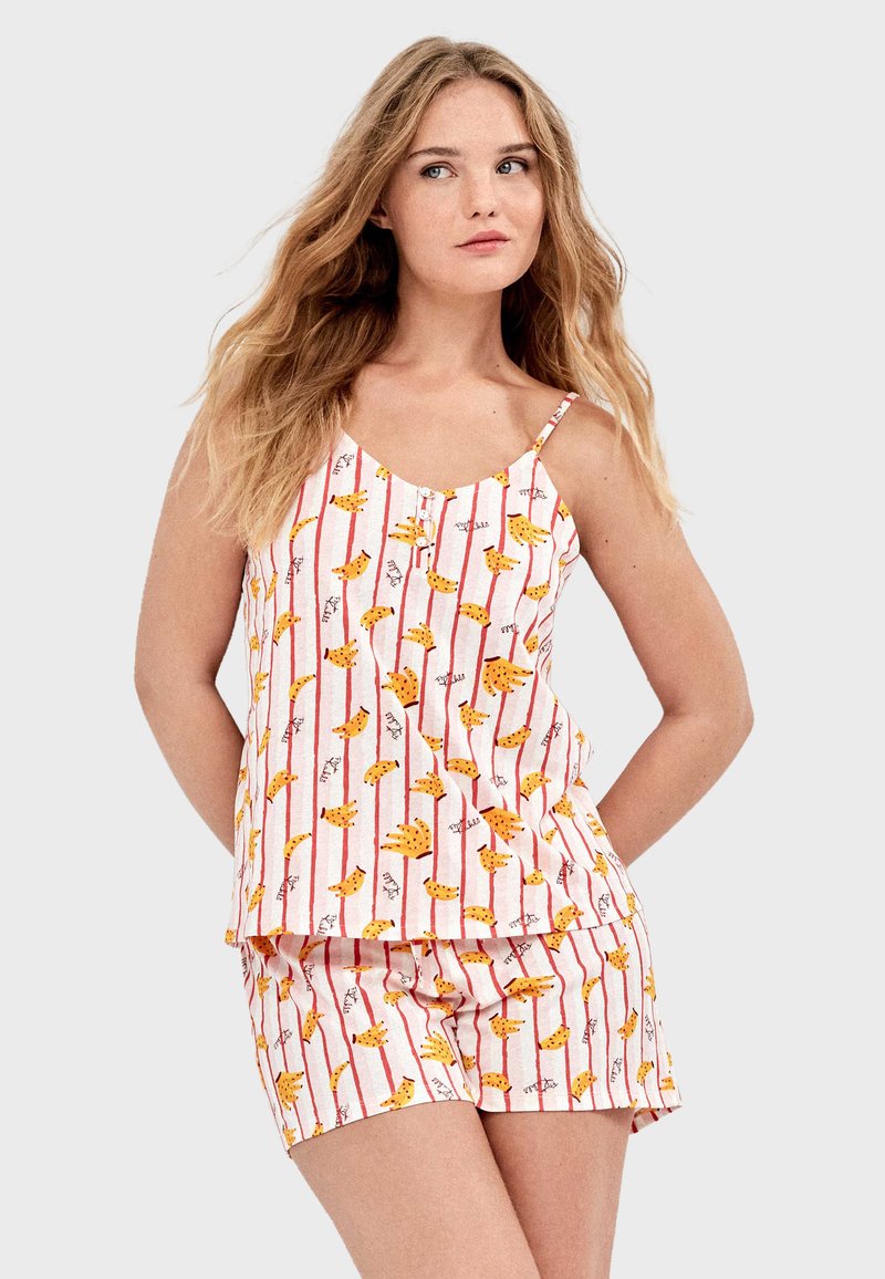Jeune femme aux cheveux blonds portant un ensemble de pyjama sans manches blanc à rayures rouges et motif floral jaune, debout les mains derrière le dos.