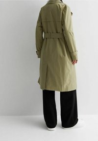 Olivově zelený trenčkot s páskem v pase, klopovými kapsami a nášivkami na ramenou, doplněný černými širokými kalhotami a bílými botami.