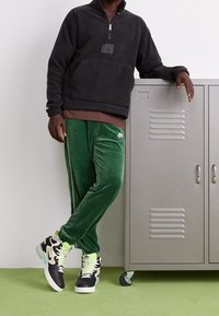 Felpa nera con zip, pantaloni di velluto verde e sneakers nere e bianche con accenti al neon, posizionati accanto a un armadietto argentato.
